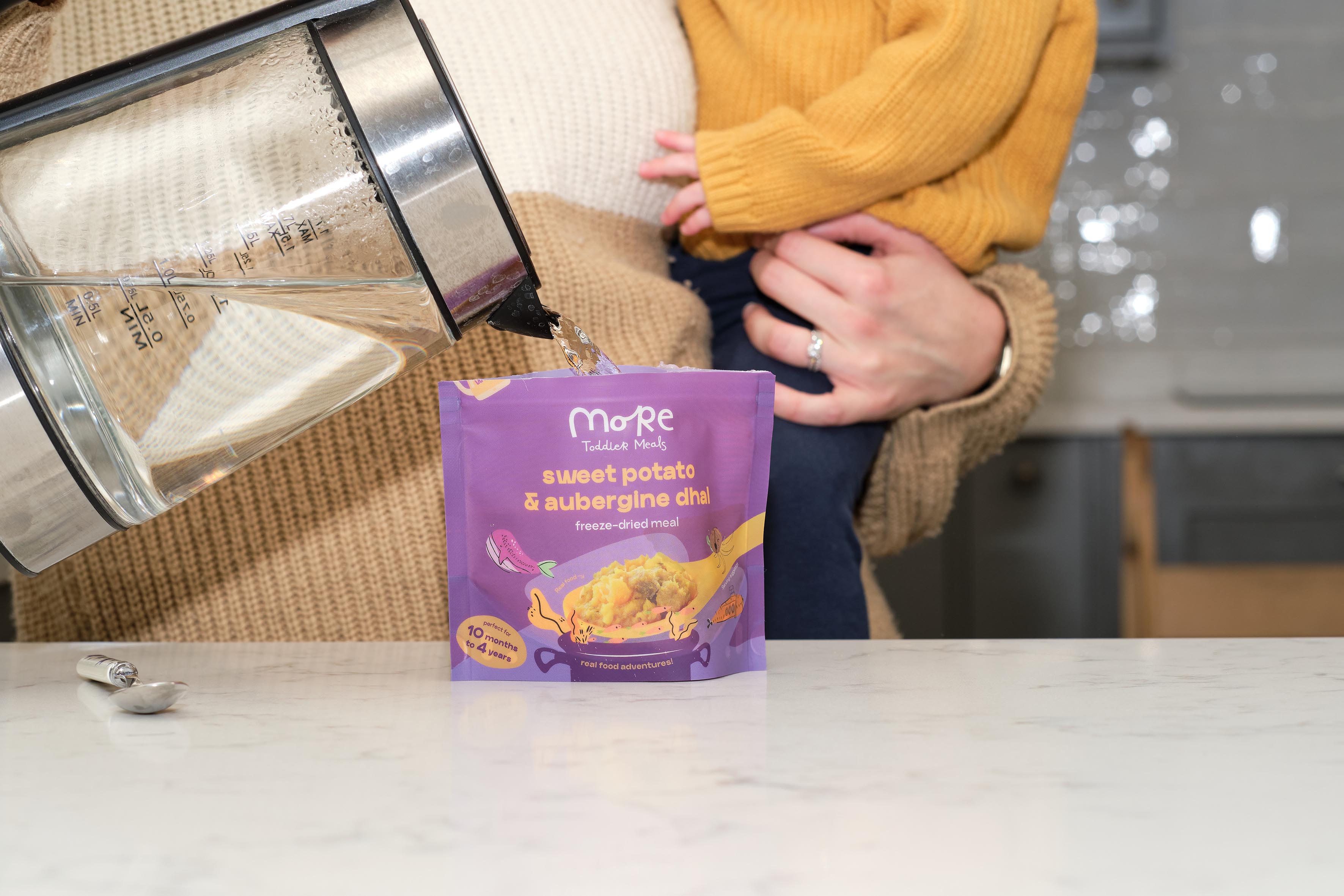 A Mum holding a toddler whilst preparing a more toddler meal with hot water. 