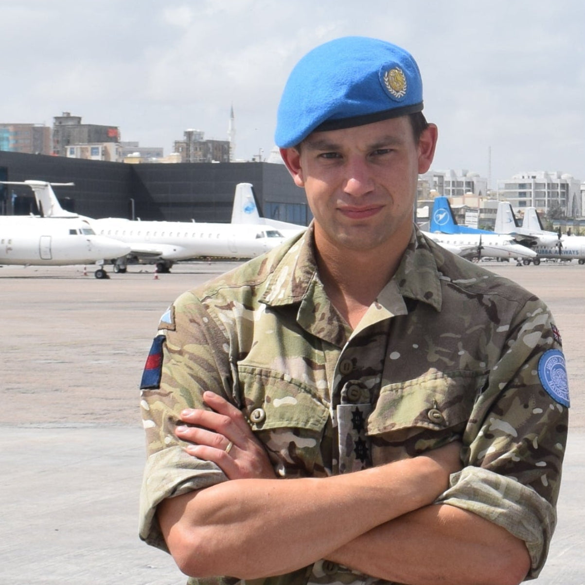 Two military officers in camouflage uniforms and blue berets standing with arms crossed on an airfield tarmac, with small aircraft and a cloudy sky in the background.